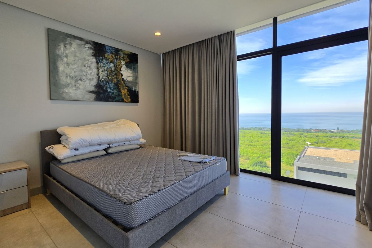 Modern bedroom featuring floor-to-ceiling windows with expansive ocean and coastal views, neutral tiled flooring, and recessed ceiling lighting.
