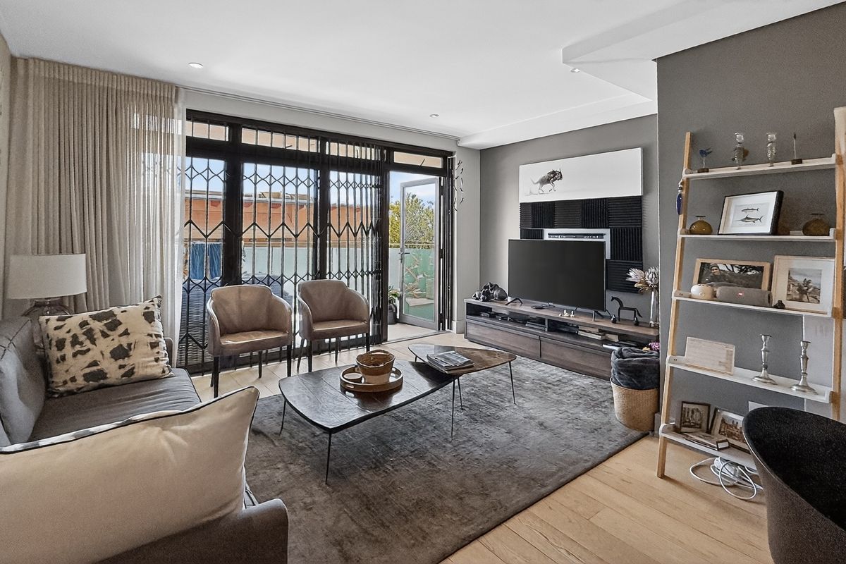 A modern, well-maintained living room featuring light wood flooring, recessed lighting, and large sliding glass doors with security gates leading to a balcony.