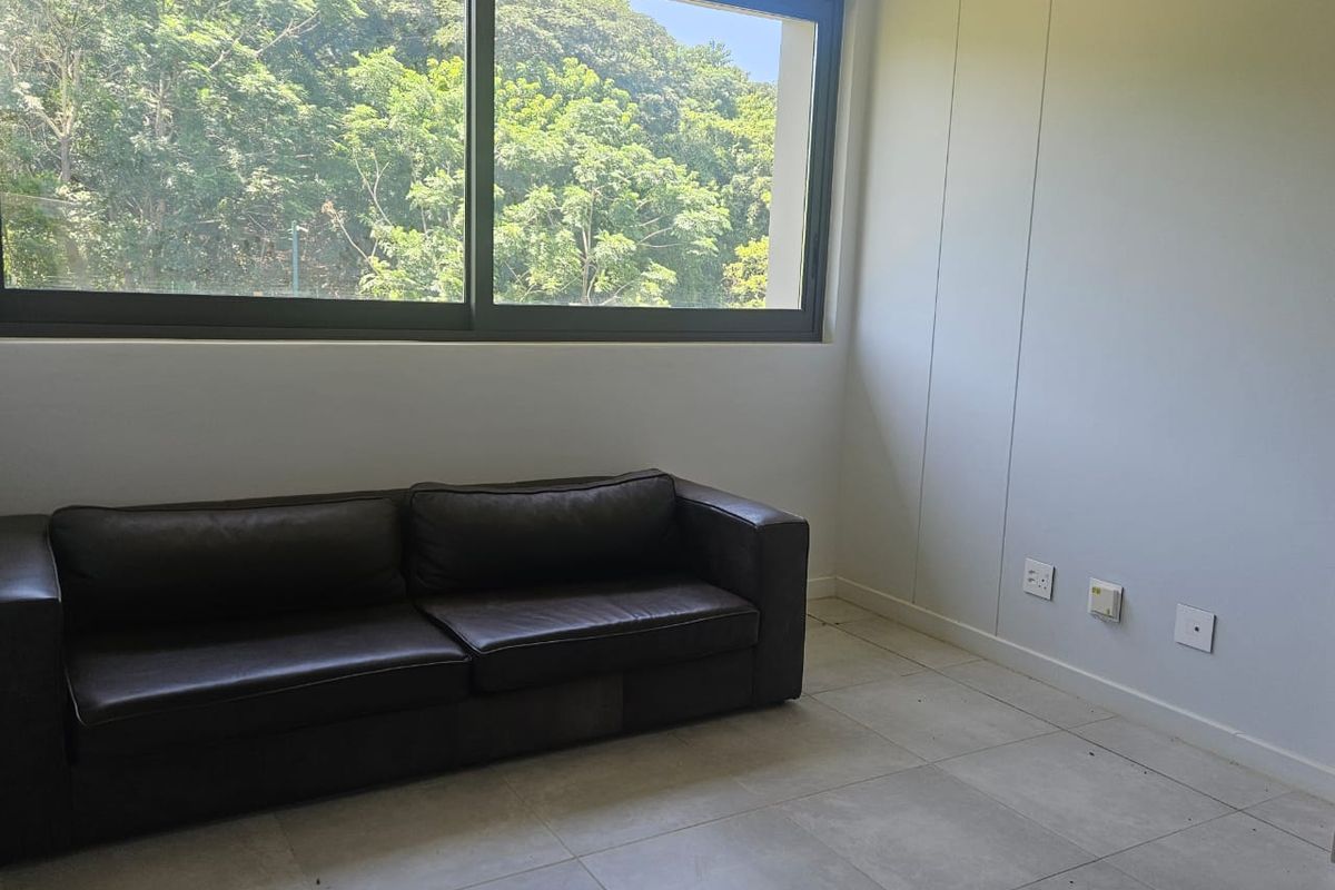 A bright living room featuring large sliding windows with a green outlook, tiled flooring, and neutral wall finishes.