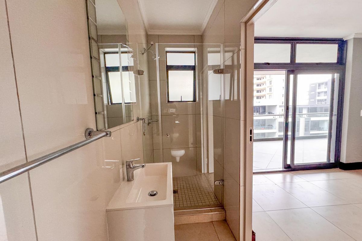 A modern, clean bathroom featuring a glass-enclosed shower, wall-mounted basin with chrome fixtures, and neutral floor-to-ceiling tiling, adjacent to an open living area.
