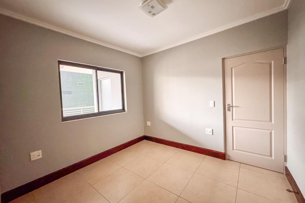 A clean, empty bedroom featuring neutral grey walls, tiled flooring, dark wood-tone skirting boards, and a sliding window for natural light.