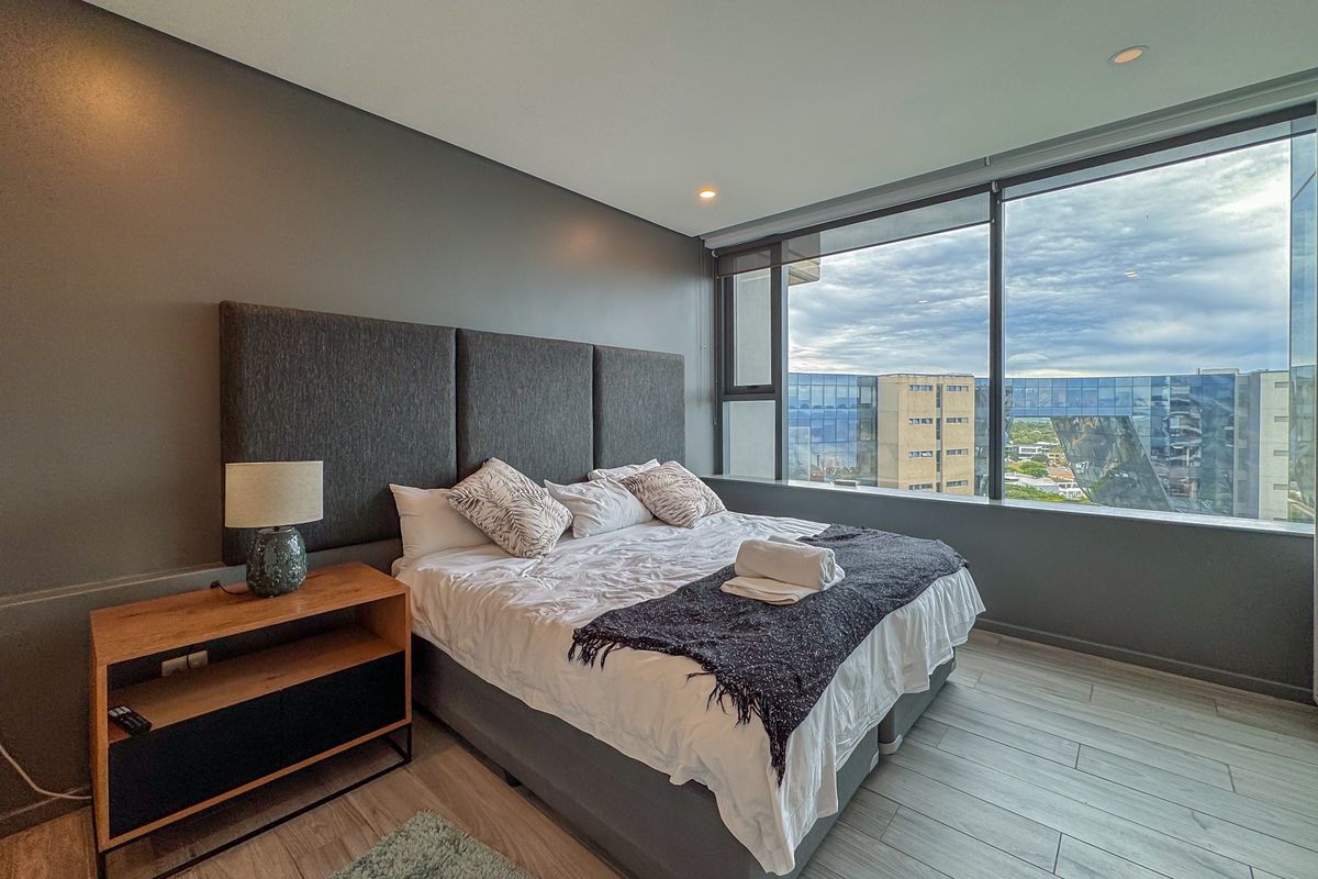 Modern bedroom featuring floor-to-ceiling windows with expansive city views, neutral grey walls, and wood-look tile flooring.
