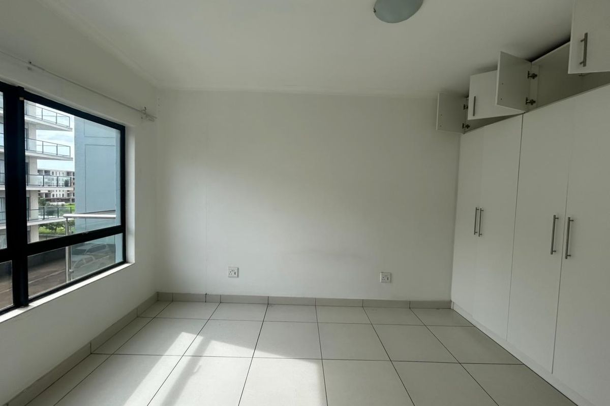 A bright, empty bedroom featuring tiled flooring, large windows, and built-in white cabinetry with overhead storage compartments.