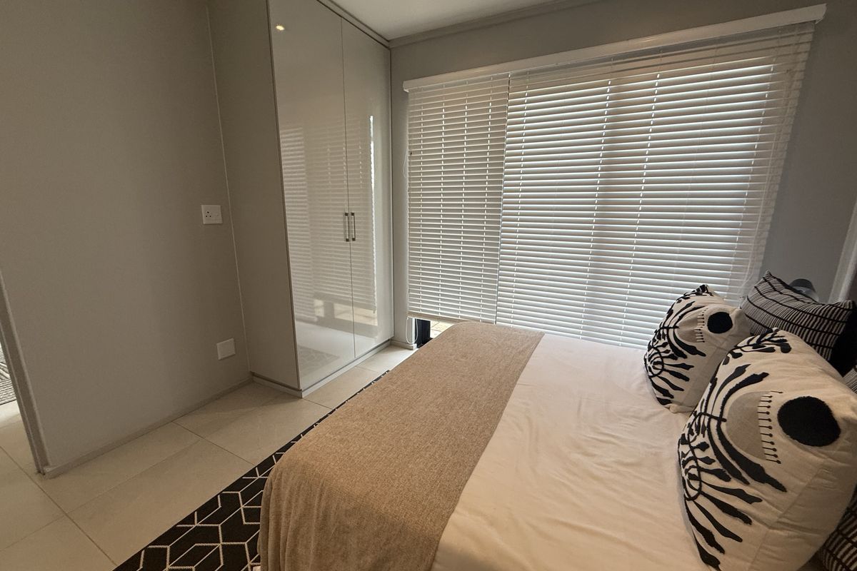 A bright bedroom featuring built-in high-gloss wardrobes, tiled flooring, and modern venetian blinds covering the window.