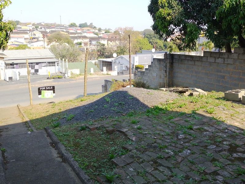 Front exterior view of the property featuring a paved driveway, concrete boundary wall, and an elevated area with loose gravel and overgrown vegetation.