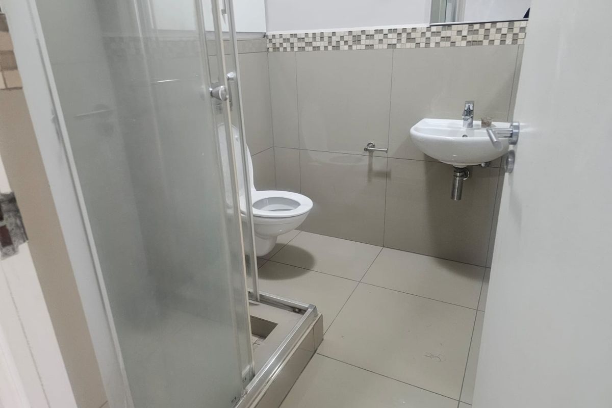 A compact bathroom featuring a glass-enclosed shower, wall-mounted basin, and toilet with neutral floor-to-ceiling tiling and a decorative mosaic border.