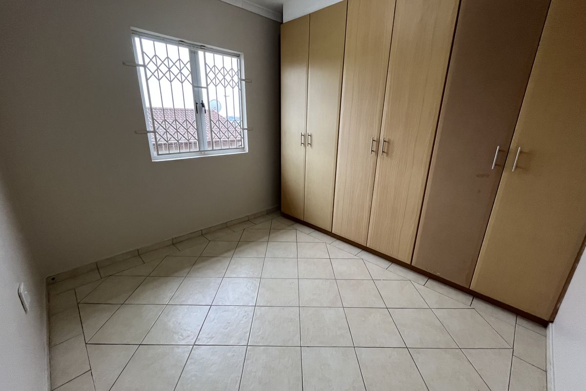A bedroom featuring tiled flooring, built-in wooden cupboards, and a window fitted with security bars.
