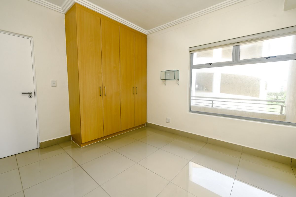 A bright, empty bedroom featuring light-toned built-in cupboards, tiled flooring, and a large window providing natural light.