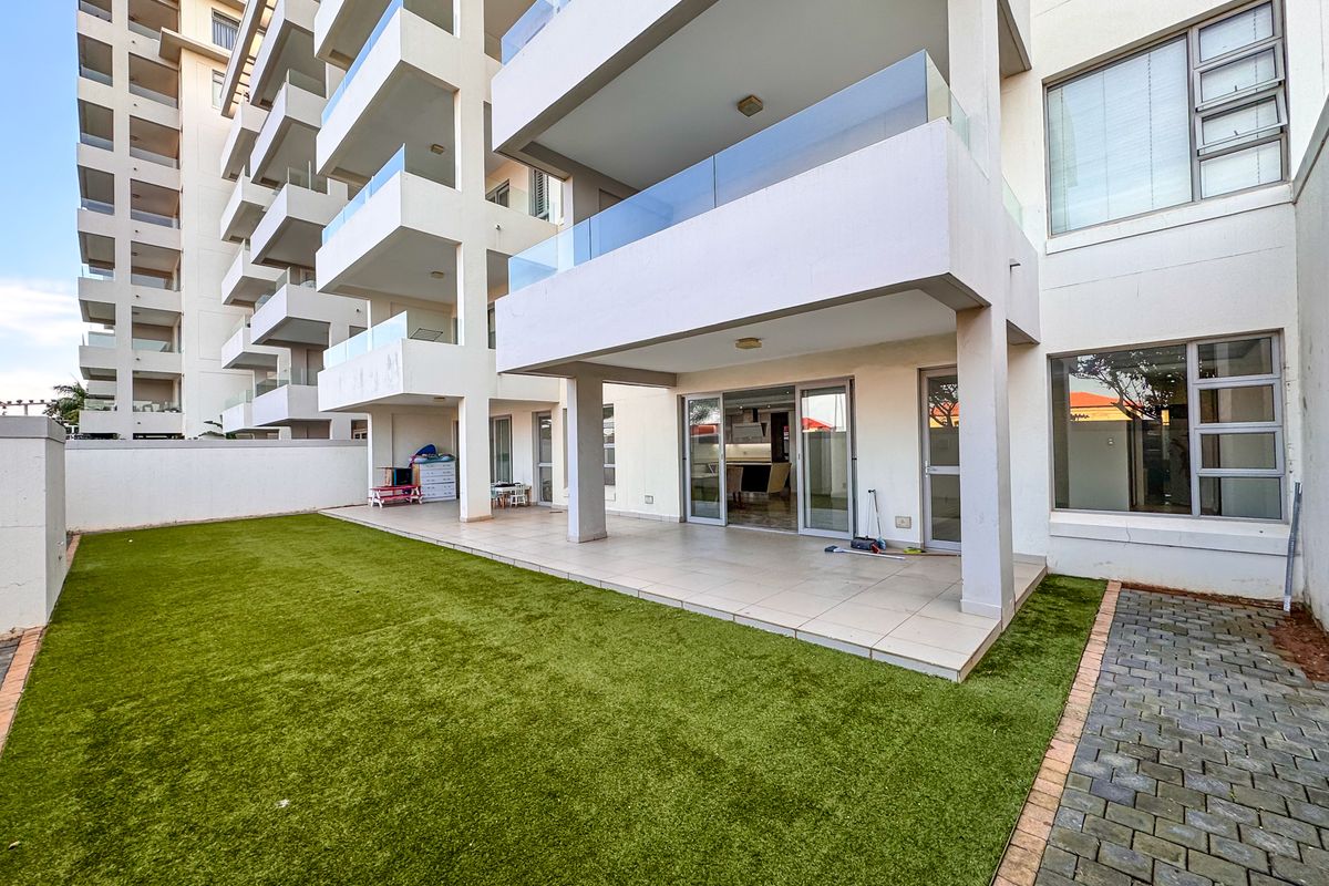 Ground floor apartment featuring a spacious covered patio with glass balustrades, leading onto a private artificial lawn garden area.