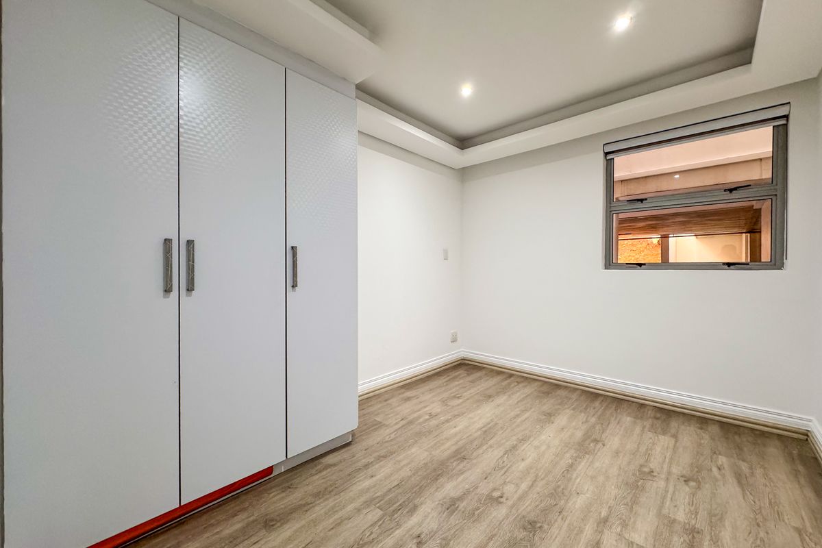 A bright, empty bedroom featuring modern wood-look flooring, recessed ceiling lighting, and built-in white cabinetry with textured doors.