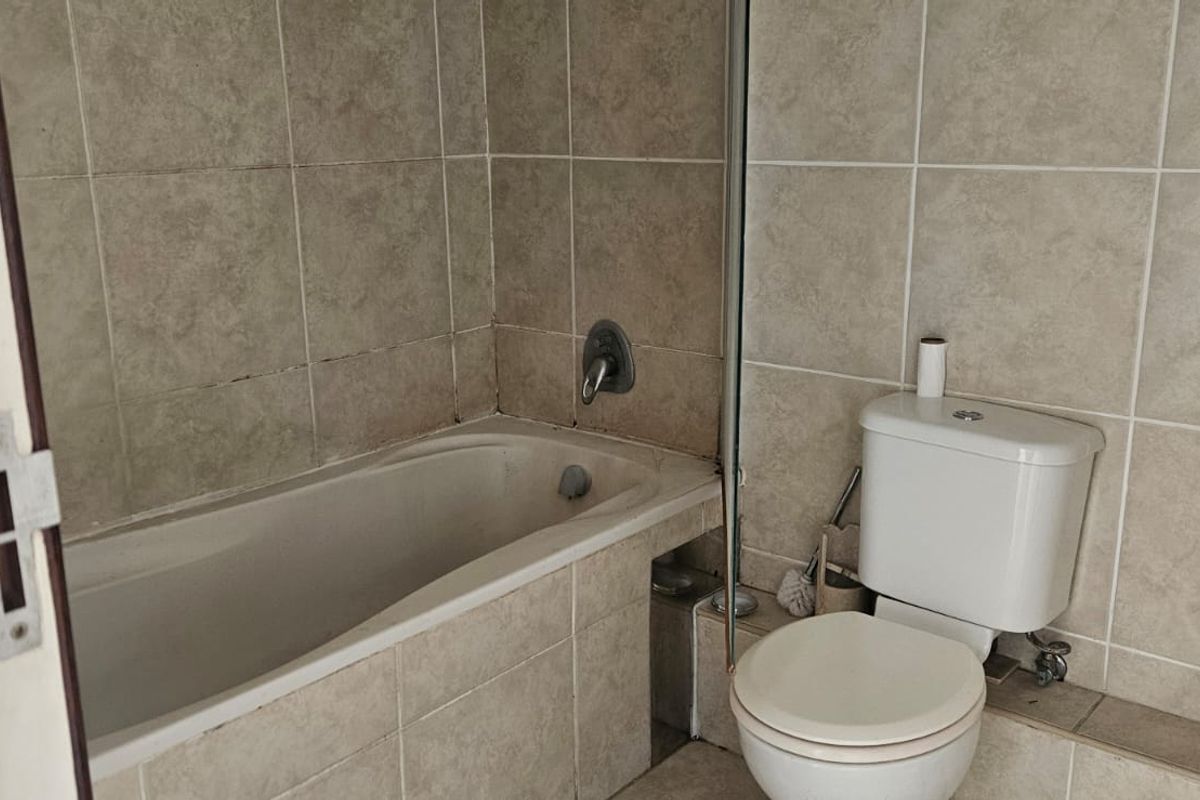 A functional bathroom featuring a built-in bathtub with shower mixer and a standard white toilet, finished with neutral wall and floor tiling.