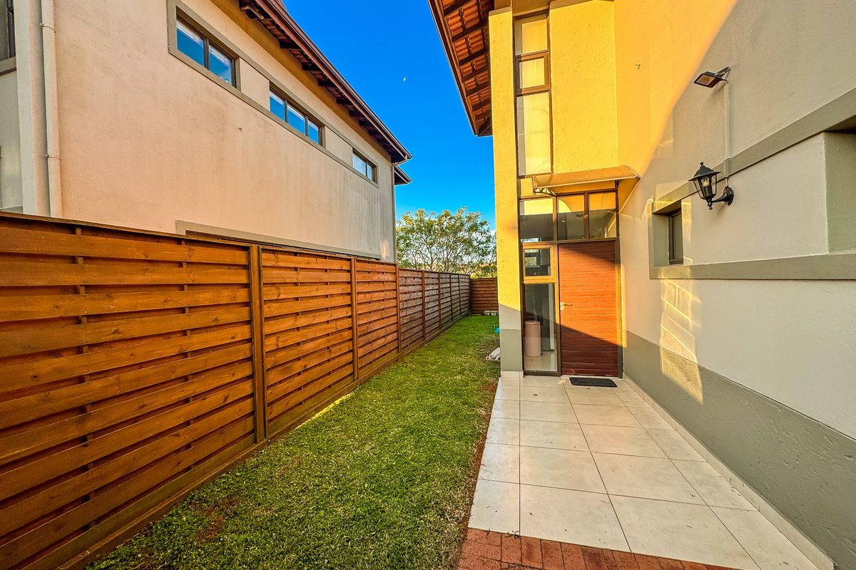 A side garden pathway featuring tiled flooring, a wooden boundary fence, and exterior wall lighting, providing access to the side entrance of the house.