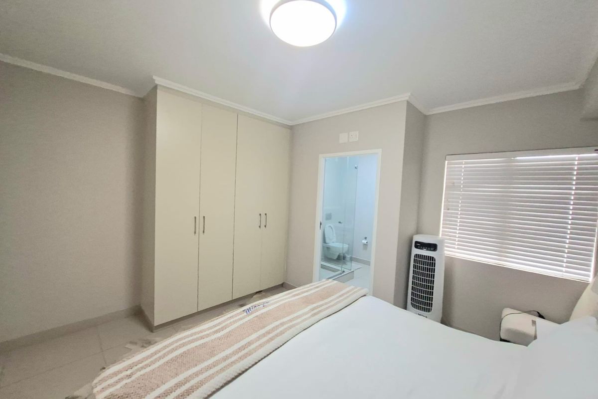 A well-maintained bedroom featuring built-in cupboards, neutral wall finishes, tiled flooring, and an en-suite bathroom with a glass door.