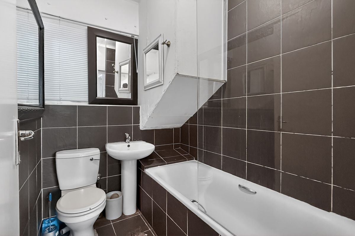 A compact bathroom featuring dark wall tiles, a white pedestal basin, a standard toilet, and a bathtub with a glass shower screen.