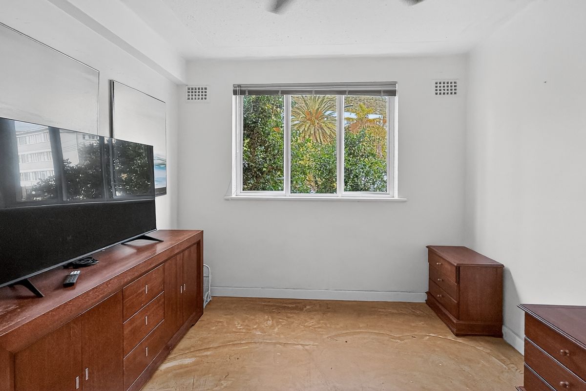 A bedroom featuring a window with garden views, ceiling fan, and plain white walls, currently awaiting new flooring and updated finishes.