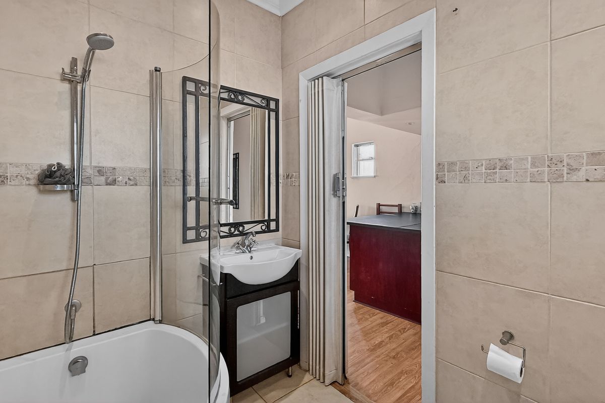 A compact bathroom featuring a shower-over-bath combination, a modern vanity unit, and neutral wall tiling, accessible via a folding door.