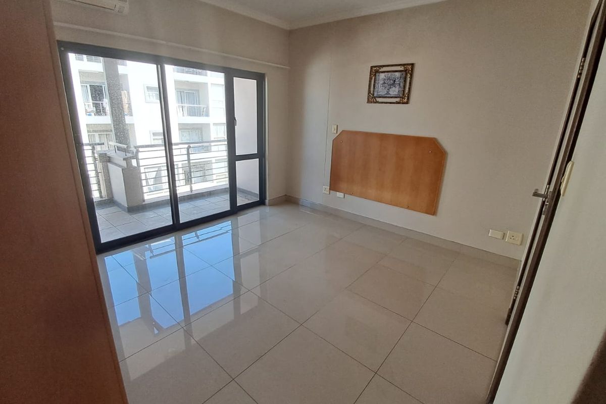 A bright, empty bedroom featuring large sliding glass doors leading to a balcony, tiled flooring, and a wall-mounted headboard panel.