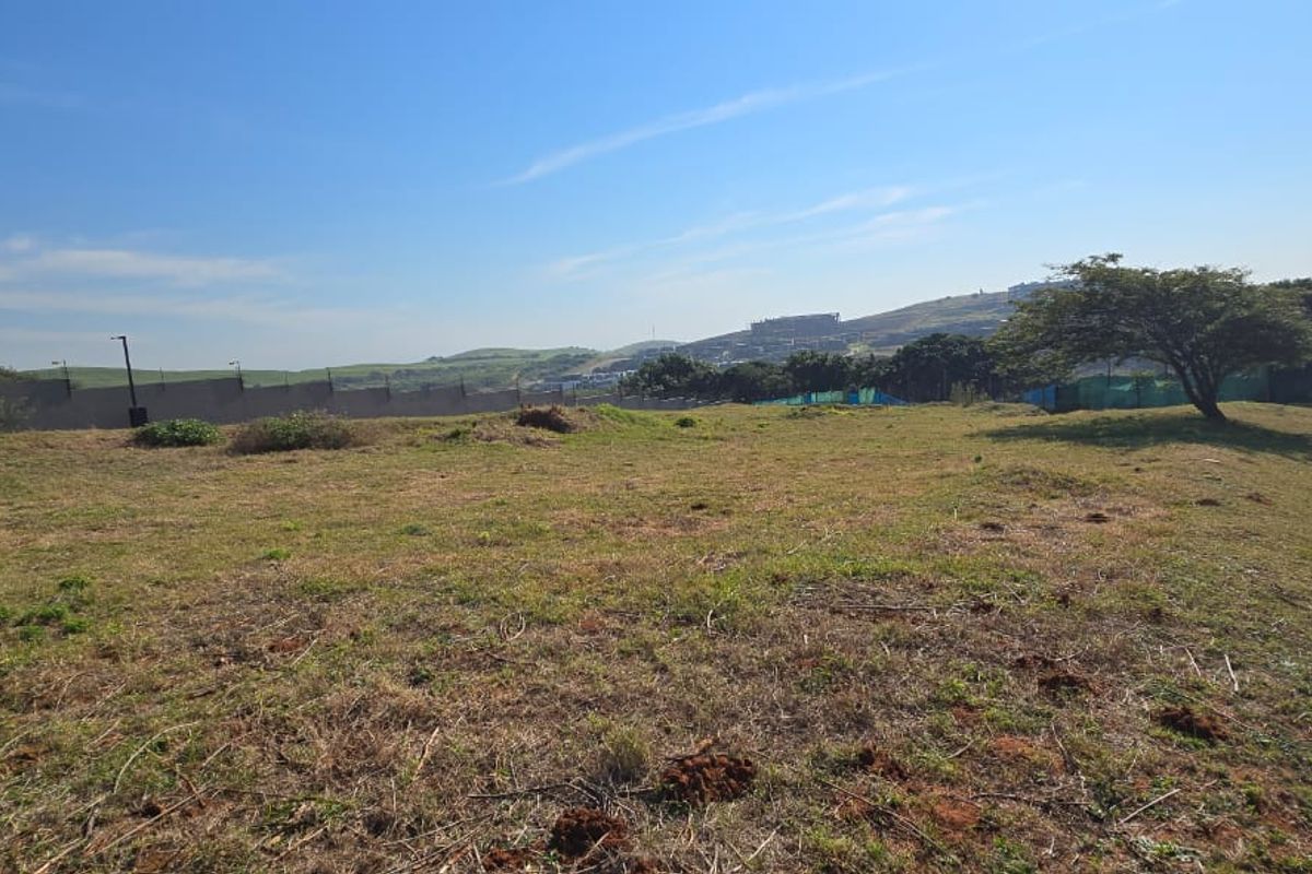 A wide-angle view of a vacant residential plot featuring natural grass terrain, perimeter boundary walls, and distant hillside views.