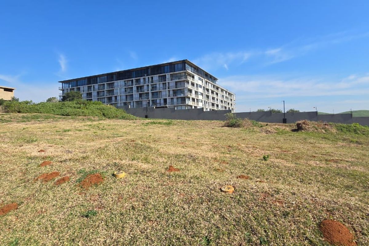 A vacant residential plot of land featuring natural grass coverage, with a view of neighboring apartment buildings and a perimeter boundary wall.