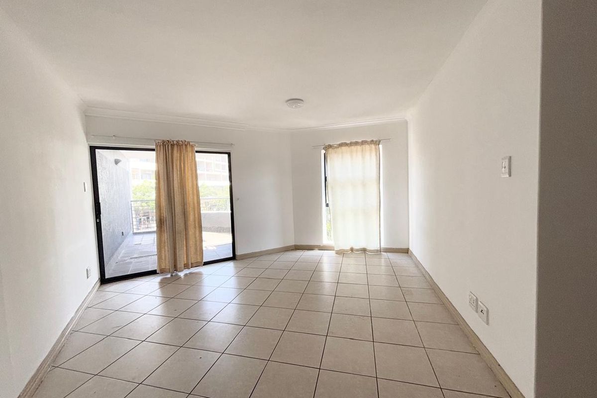 An empty, open-plan living area featuring neutral tiled flooring, cove cornicing, and a sliding glass door providing access to an outdoor balcony.