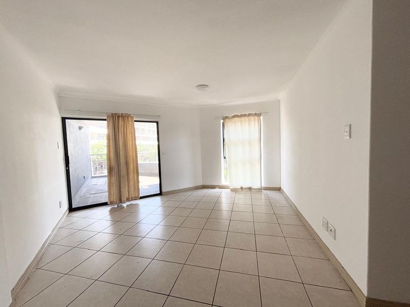 An empty, open-plan living area featuring neutral tiled flooring, cove cornicing, and a sliding glass door providing access to an outdoor balcony.