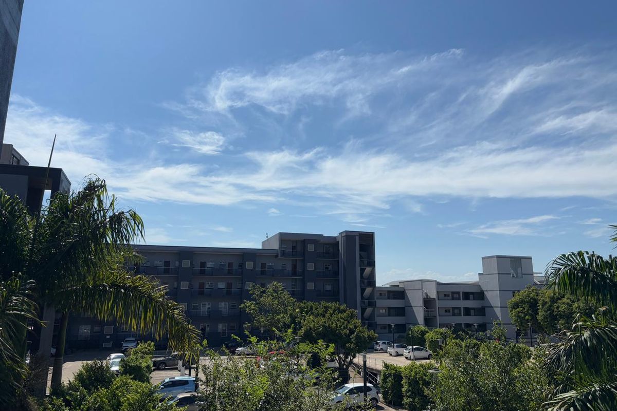 An elevated view of a multi-story apartment complex featuring a paved parking area and surrounding mature trees under a clear blue sky.