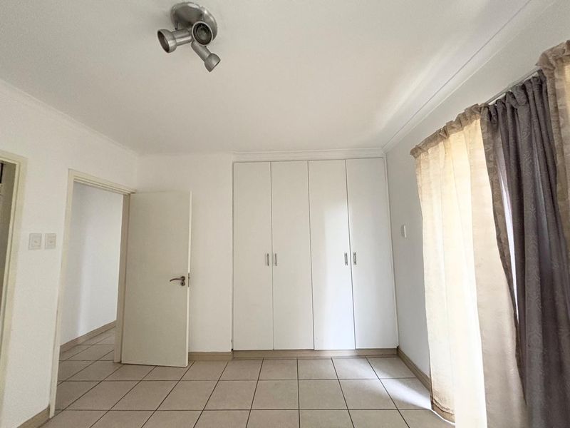 A bright, empty bedroom featuring neutral tiled flooring, built-in white cupboards, and a modern ceiling spotlight fixture.