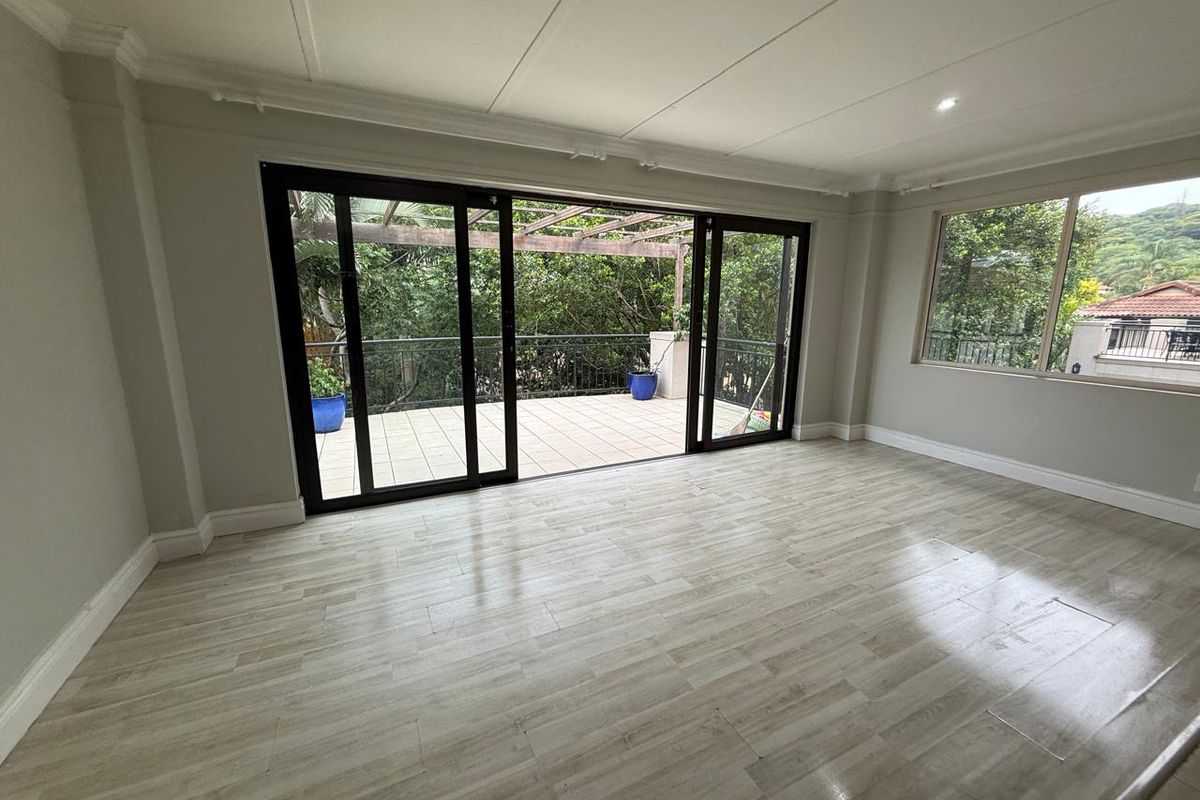 Bright living area featuring light wood-look flooring, recessed ceiling lighting, and large sliding glass doors opening onto a private balcony with a pergola.