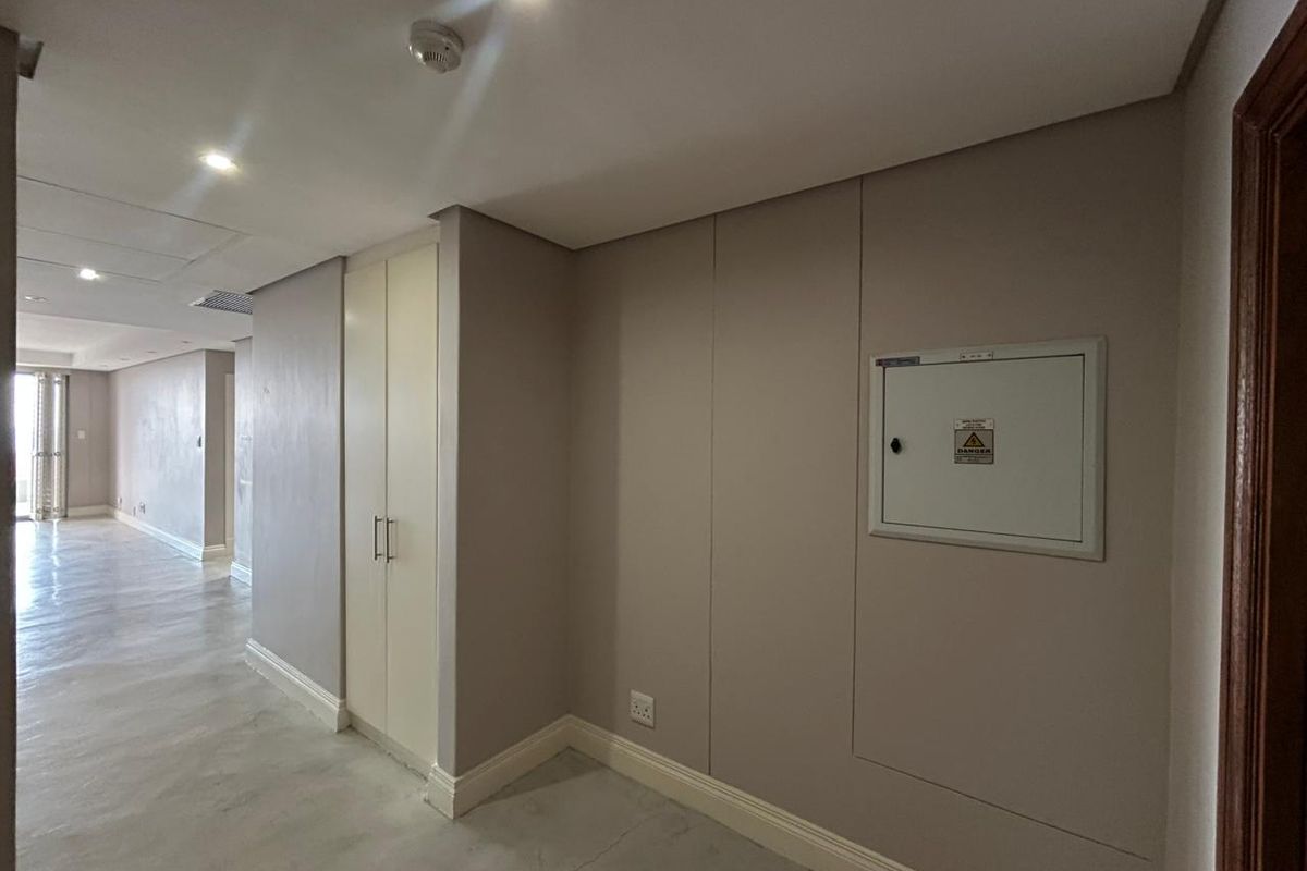 A clean, neutral hallway featuring recessed downlighting, a built-in storage cupboard, and a wall-mounted electrical distribution board.