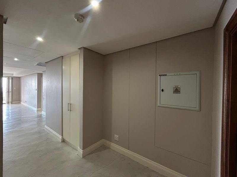 A clean, neutral hallway featuring recessed downlighting, a built-in storage cupboard, and a wall-mounted electrical distribution board.