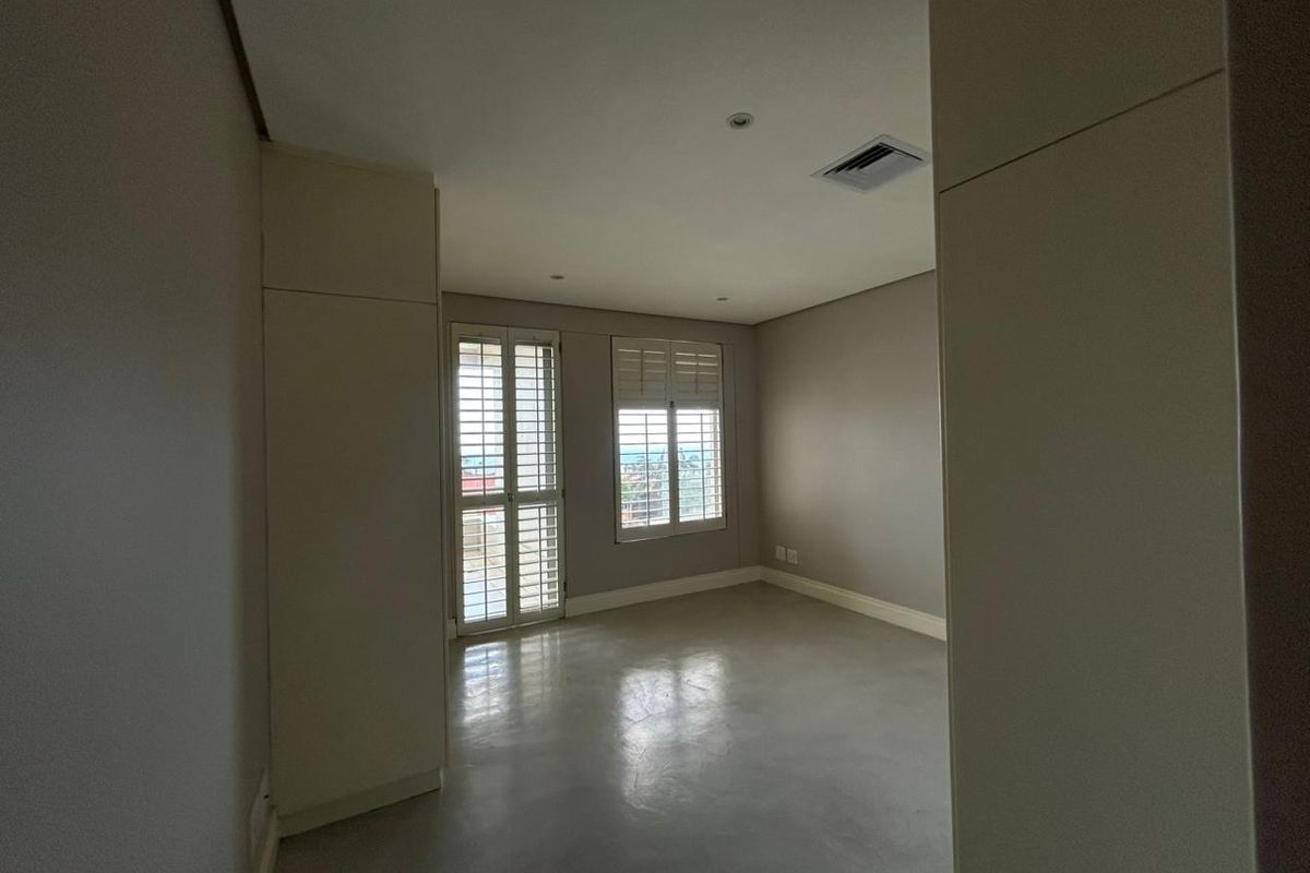 A spacious, empty bedroom featuring neutral walls, polished concrete flooring, built-in cupboards, and white plantation shutters allowing natural light.