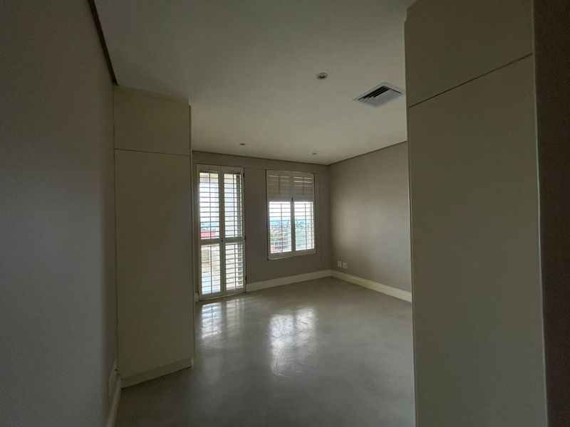 A spacious, empty bedroom featuring neutral walls, polished concrete flooring, built-in cupboards, and white plantation shutters allowing natural light.