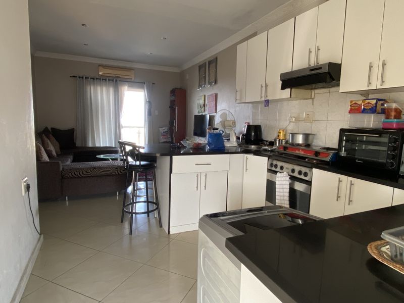 Open-plan kitchen and living area featuring white cabinetry, dark countertops, tiled flooring, and an integrated air conditioning unit leading to a balcony.