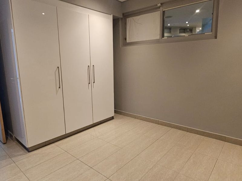 A clean, empty bedroom featuring modern white built-in cupboards, neutral tiled flooring, and a high-level window, offering a bright and versatile living space.