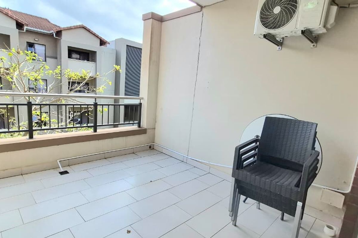 A spacious apartment balcony featuring tiled flooring, a secure metal balustrade, and a wall-mounted air conditioning unit, offering a pleasant outdoor area.