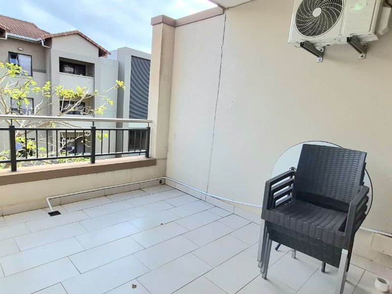 A spacious apartment balcony featuring tiled flooring, a secure metal balustrade, and a wall-mounted air conditioning unit, offering a pleasant outdoor area.