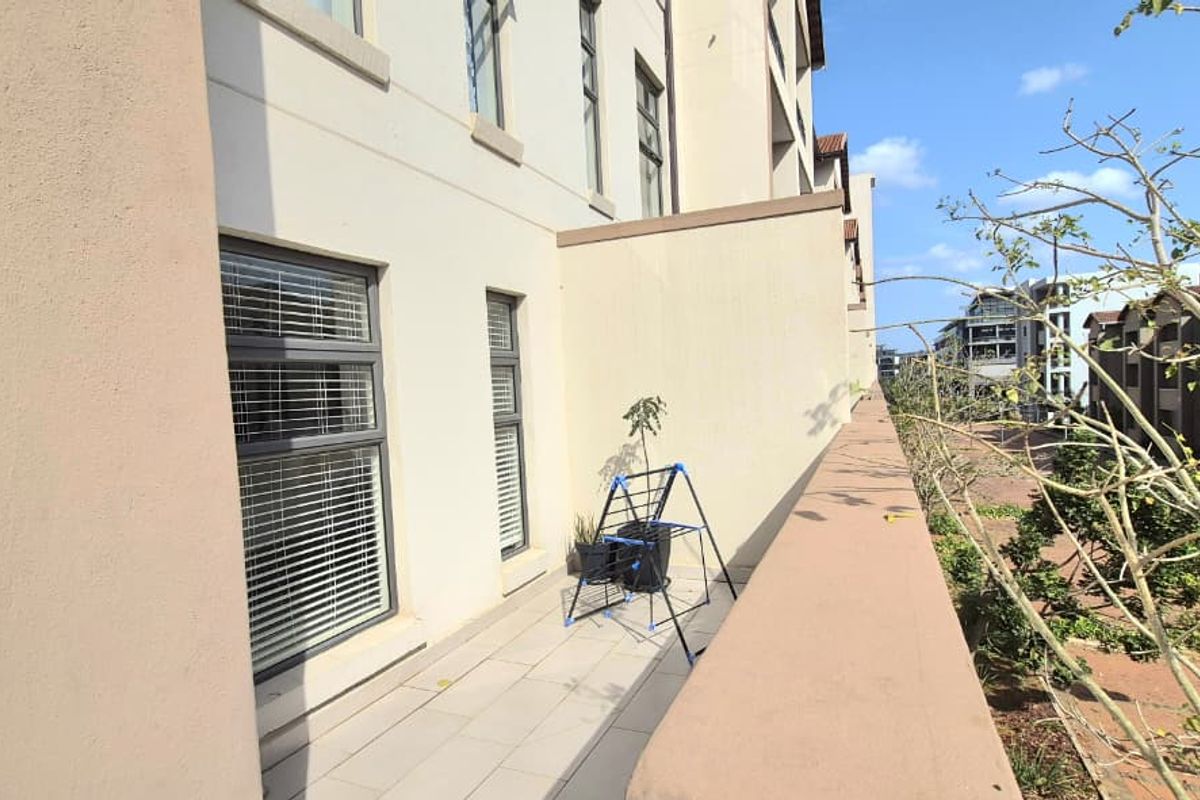 A spacious apartment balcony featuring tiled flooring, aluminium window frames, and a solid balustrade overlooking the complex grounds.