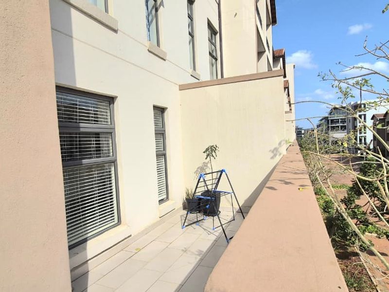 A spacious apartment balcony featuring tiled flooring, aluminium window frames, and a solid balustrade overlooking the complex grounds.