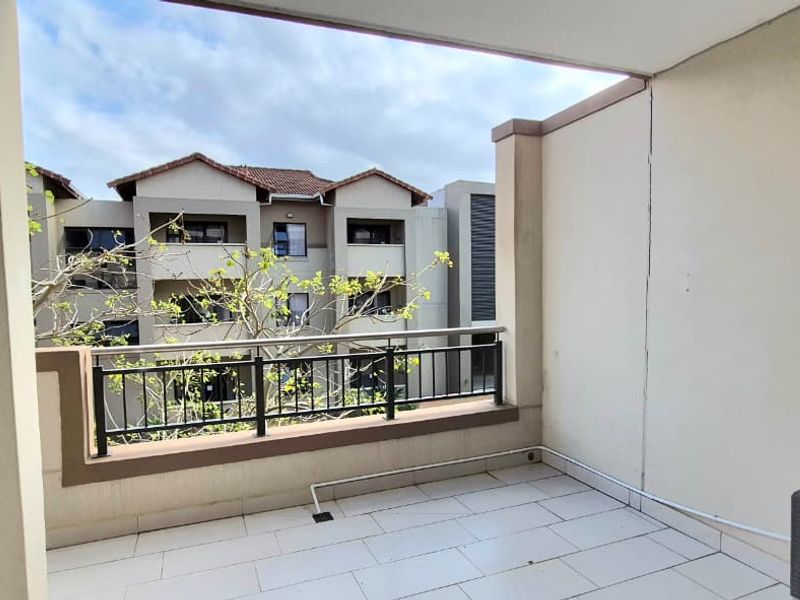 A covered balcony featuring tiled flooring, a metal balustrade, and a wall-mounted air conditioning unit, overlooking the apartment complex grounds.