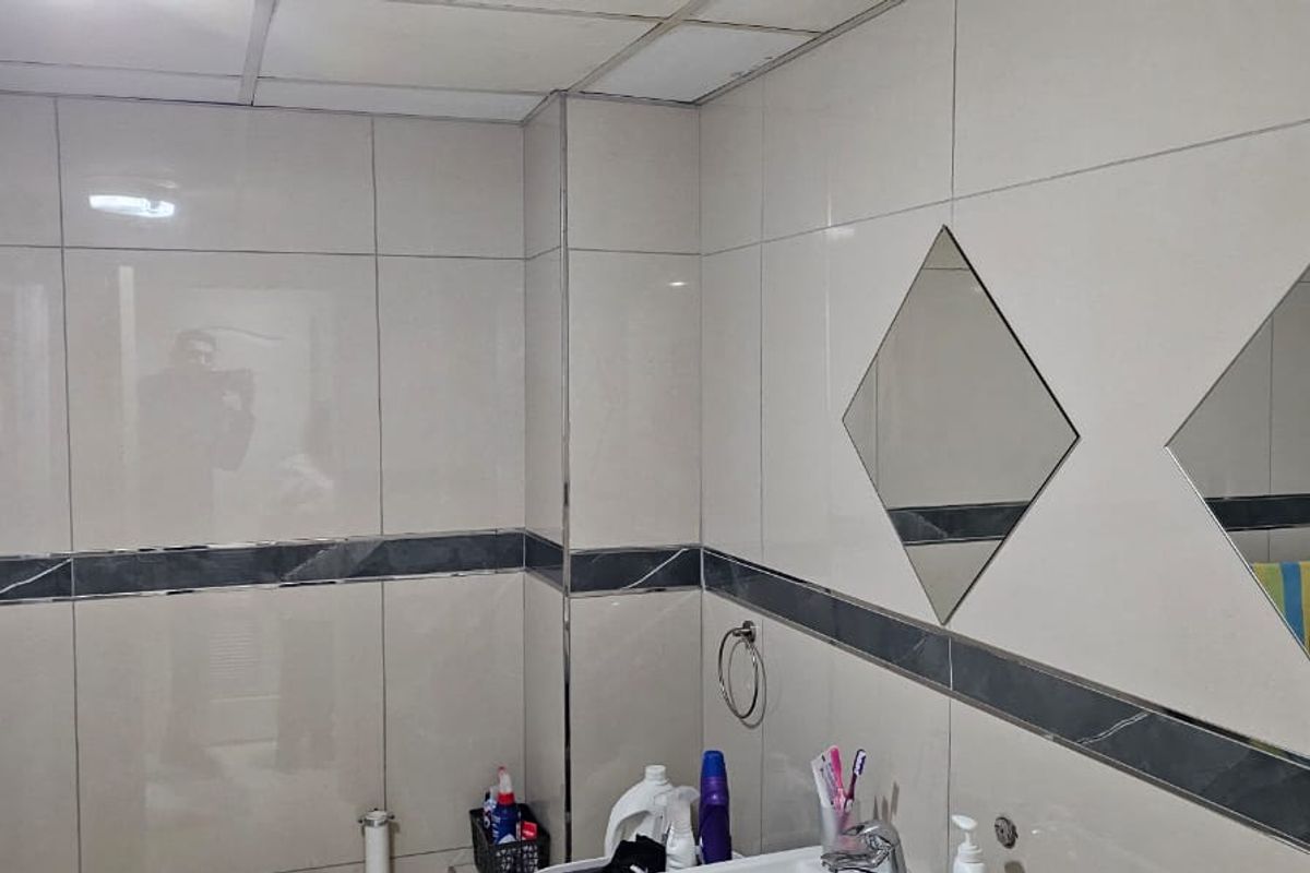 A functional bathroom featuring tiled walls with a decorative border, drop ceiling, and wall-mounted diamond-shaped mirrors.