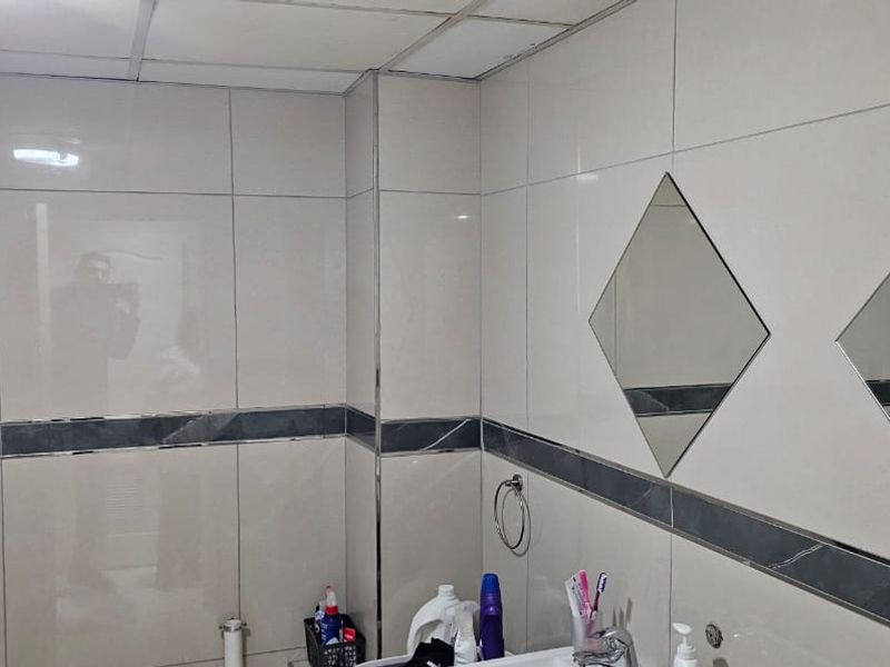 A functional bathroom featuring tiled walls with a decorative border, drop ceiling, and wall-mounted diamond-shaped mirrors.