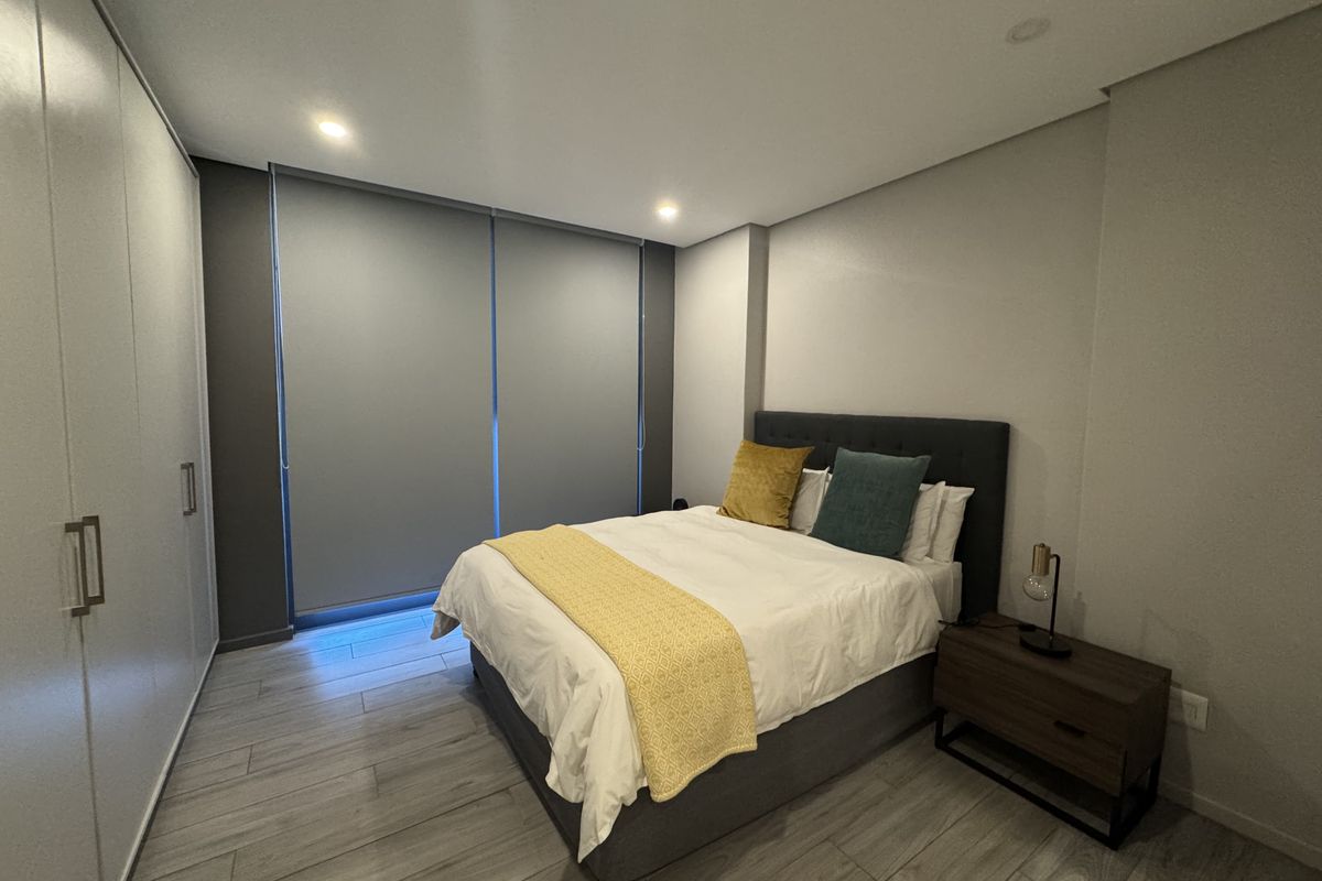 Modern bedroom featuring built-in cupboards, neutral tiled flooring, recessed lighting, and large roller blinds for privacy.