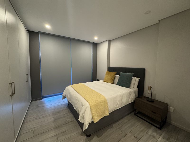 Modern bedroom featuring built-in cupboards, neutral tiled flooring, recessed lighting, and large roller blinds for privacy.