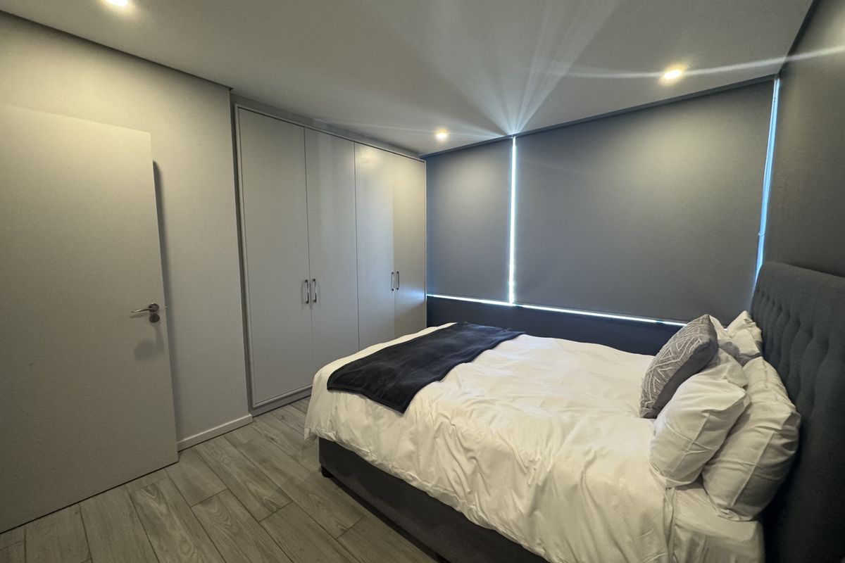 A modern, well-maintained bedroom featuring neutral tones, built-in cupboards, recessed lighting, and tiled flooring with roller blinds for privacy.