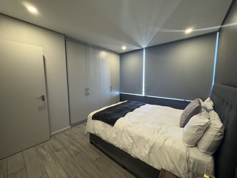 A modern, well-maintained bedroom featuring neutral tones, built-in cupboards, recessed lighting, and tiled flooring with roller blinds for privacy.