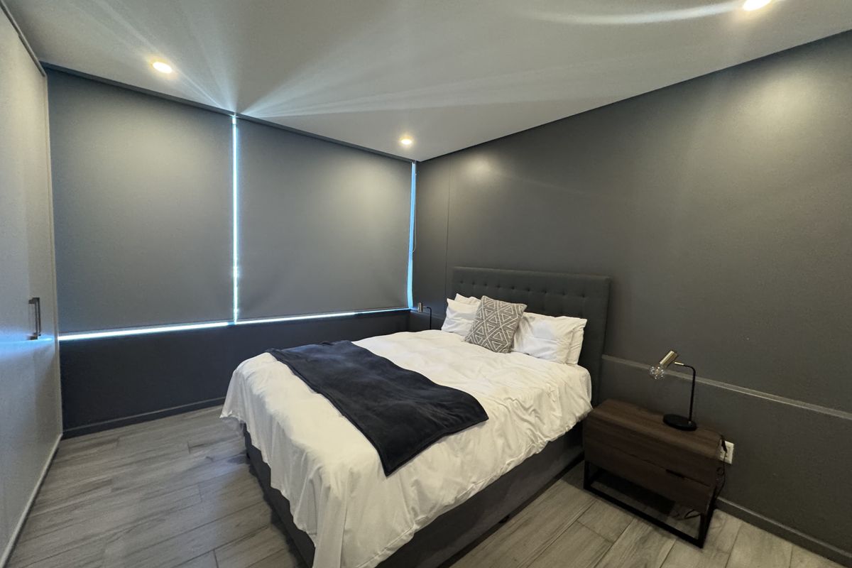 A modern bedroom featuring dark grey walls, wood-look tile flooring, recessed downlighting, and integrated roller blinds for privacy.