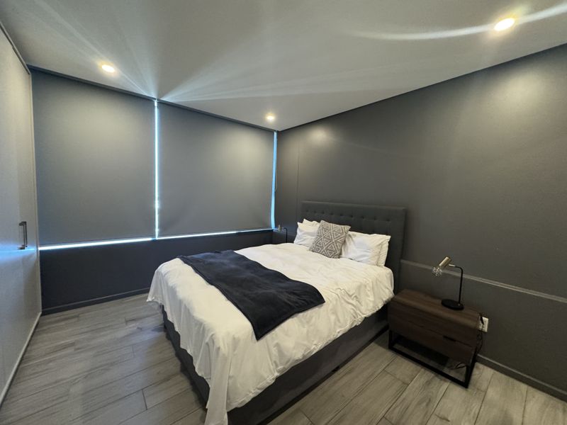 A modern bedroom featuring dark grey walls, wood-look tile flooring, recessed downlighting, and integrated roller blinds for privacy.