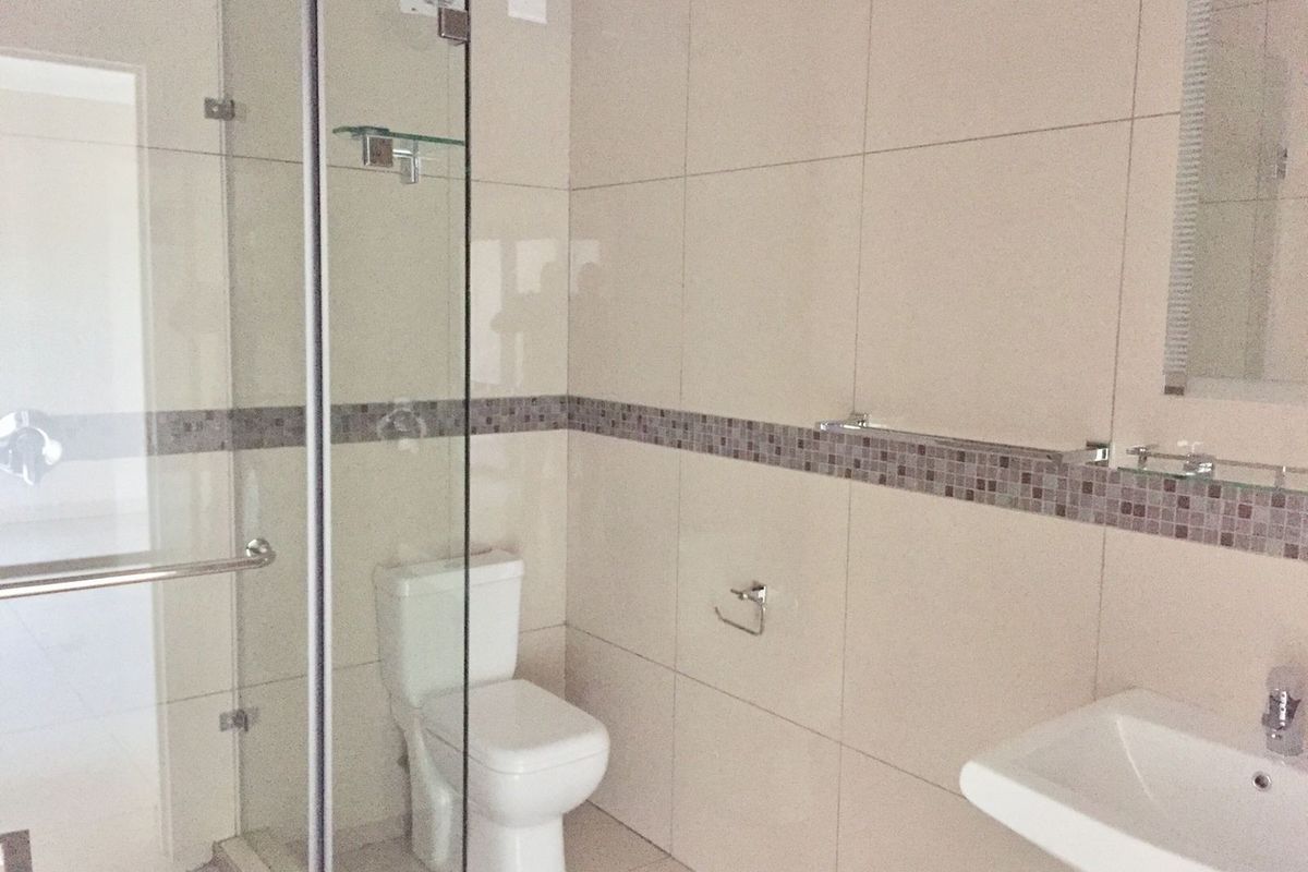 A clean, modern bathroom featuring a glass-enclosed shower, pedestal basin, and neutral wall tiling with a decorative mosaic accent border.