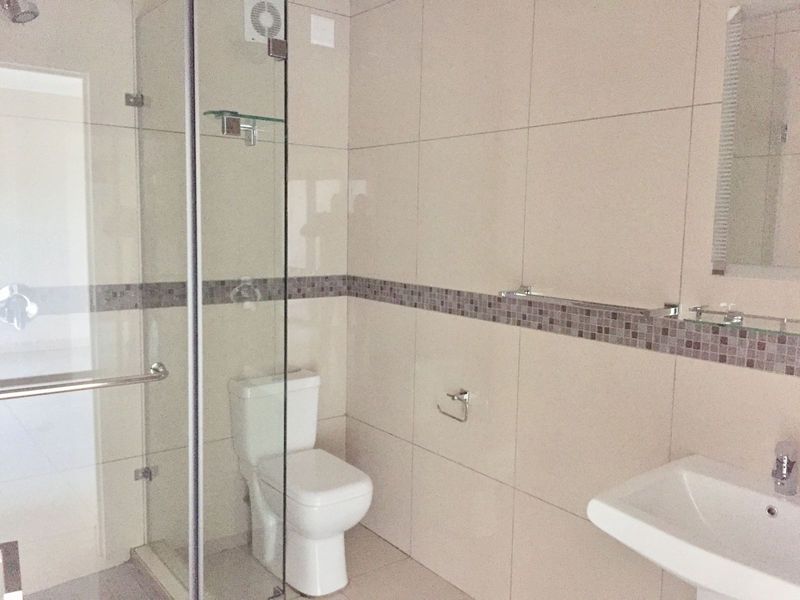 A clean, modern bathroom featuring a glass-enclosed shower, pedestal basin, and neutral wall tiling with a decorative mosaic accent border.