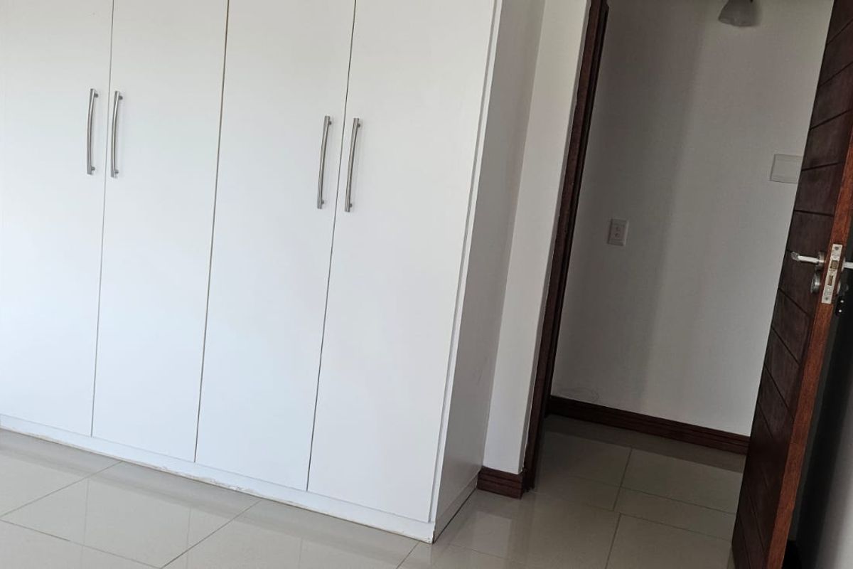 A clean, empty bedroom featuring white built-in cupboards, neutral tiled flooring, and a dark wood internal door.